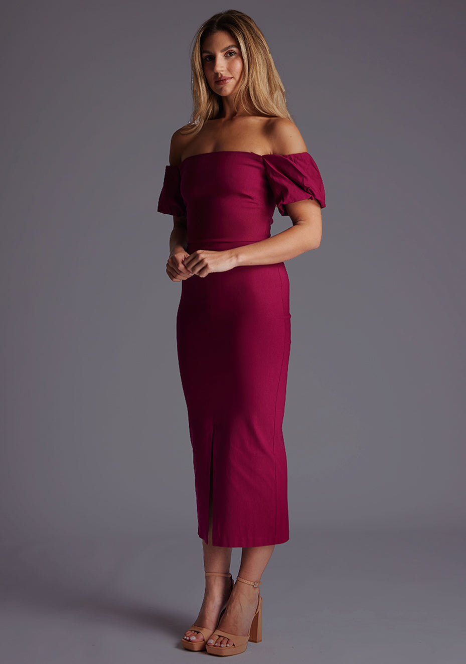 Front quarter image of a blonde model wearing a berry midaxi dress with a bardot neckline gracefully frame off shoulders, while the back zip ensures a seamless silhouette. The design featured is the Vesper Quinton Berry Midaxi Dress.