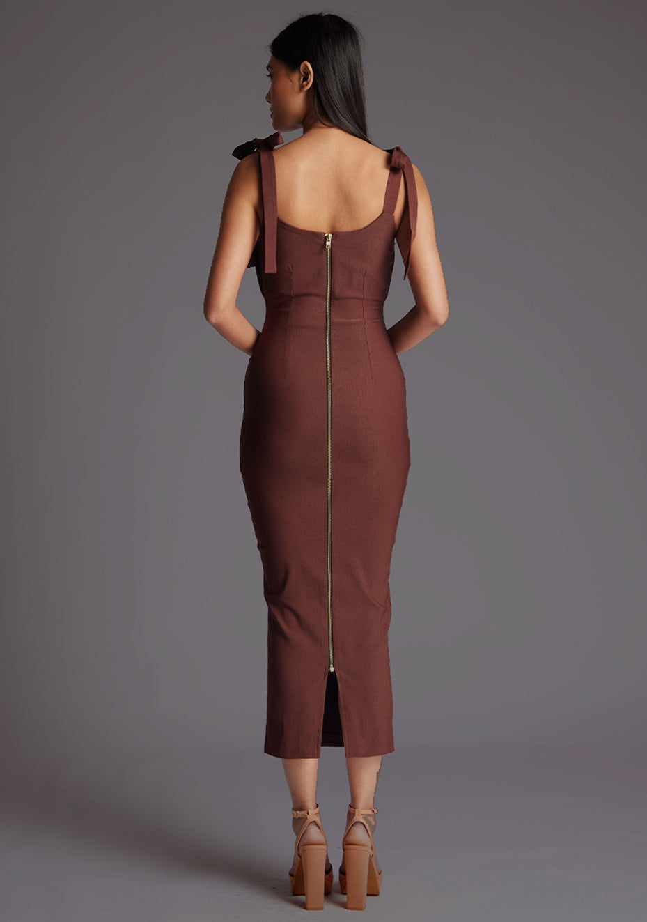 Back image of a model wearing a chocolate midaxi dress with a subtle sweetheart neckline, self-tie bow straps, pleating detail at bust, bodycon fit with statement gold zip at centre back .The design featured is the Vesper Rapunzel Chocolate Midaxi Dress.