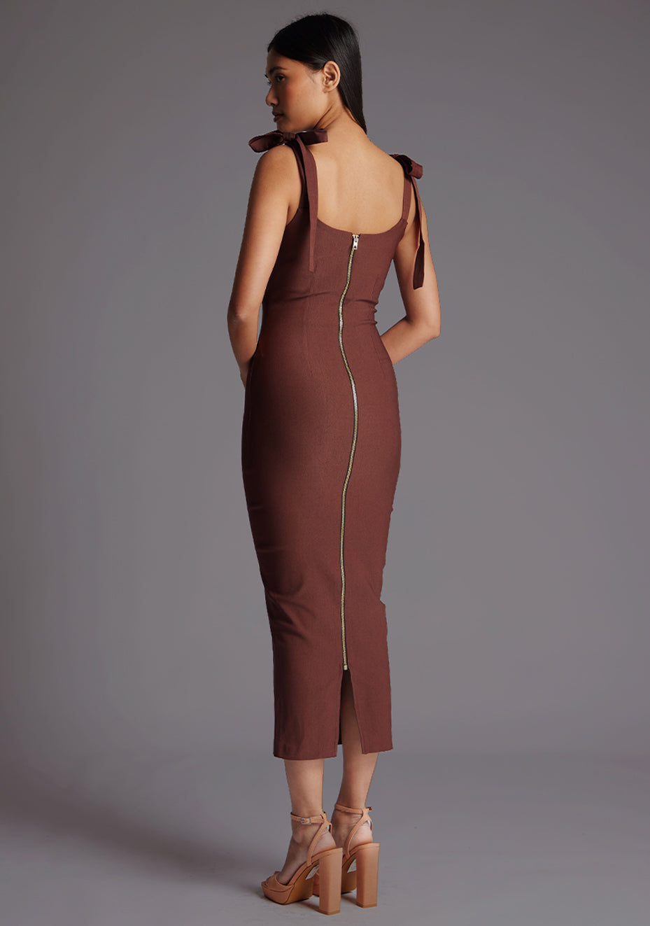 Back quarter image of a model wearing a chocolate midaxi dress with a subtle sweetheart neckline, self-tie bow straps, pleating detail at bust, bodycon fit with statement gold zip at centre back .The design featured is the Vesper Rapunzel Chocolate Midaxi Dress.