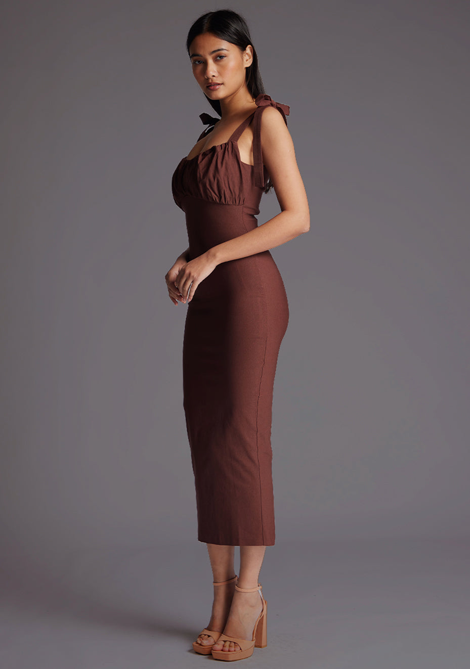 Front quarter image of a model wearing a chocolate midaxi dress with a subtle sweetheart neckline, self-tie bow straps, pleating detail at bust, bodycon fit with statement gold zip at centre back .The design featured is the Vesper Rapunzel Chocolate Midaxi Dress.