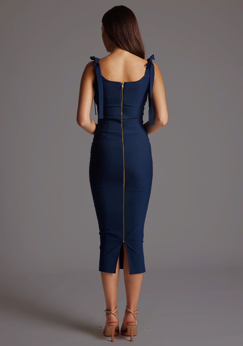 Back image of a model wearing a navy bow midaxi dress with a subtle sweetheart neckline, self-tie bow straps, pleating detail at bust, bodycon fit with statement gold zip at centre back .The design featured is the Vesper Rapunzel Navy Bow Midaxi Dress.