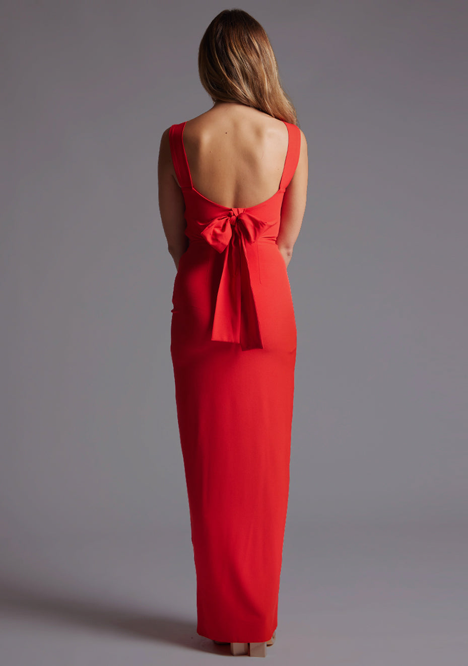 Back image of a blonde model wearing a red bow back maxi dress with a v neckline adds a touch of allure, front skirt split, while the bodycon fit flatters your figure elegantly and adding a touch of glamour. The design featured is the Vesper Reese Red Bow Back Maxi Dress