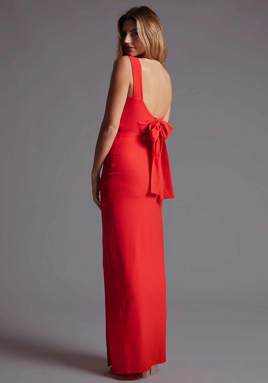 Back quarter image of a blonde model wearing a red bow back maxi dress with a v neckline adds a touch of allure, front skirt split, while the bodycon fit flatters your figure elegantly and adding a touch of glamour. The design featured is the Vesper Reese Red Bow Back Maxi Dress