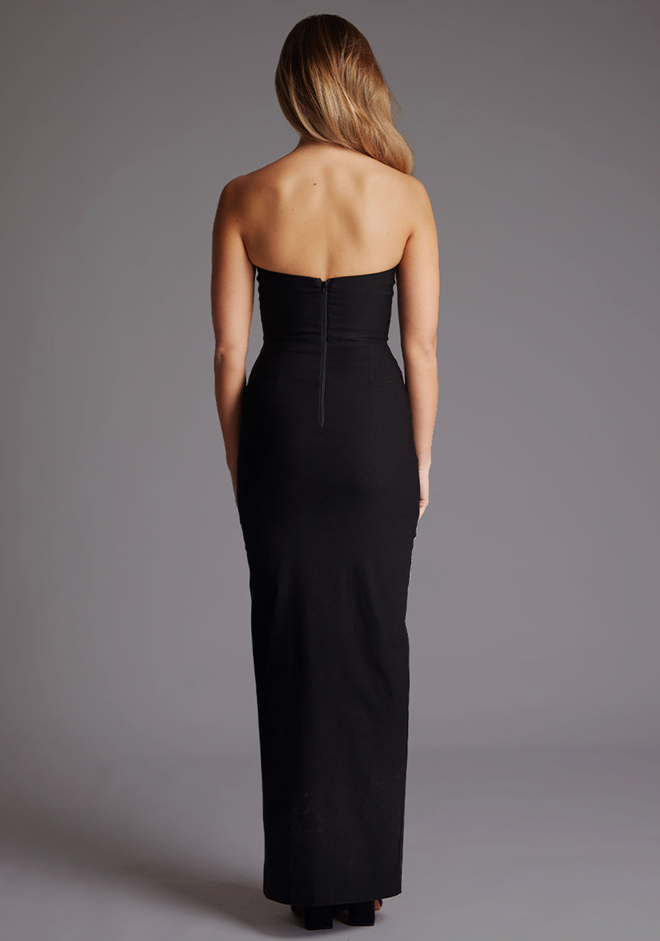 Back image of a blonde model wearing a black bodycon maxi dress with a  strapless elegance with a front skirt split for graceful movement and flattering slim fit enhances silhouette.  The design featured is the Vesper Remington Black Bodycon Maxi Dress.