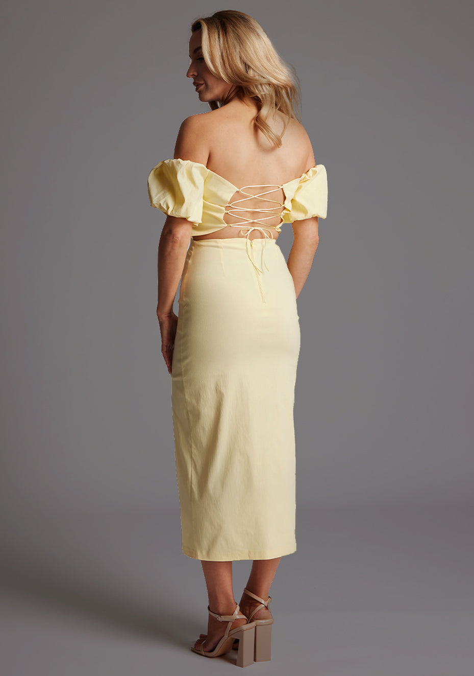 Back quarter image of a blonde model wearing a yellow bardot midaxi dress with a subtle sweetheart neckline, statement puff sleeves, a front skirt split and lace-up detail at the back. The design featured is the Vesper River Yellow Bardot Midaxi Dress