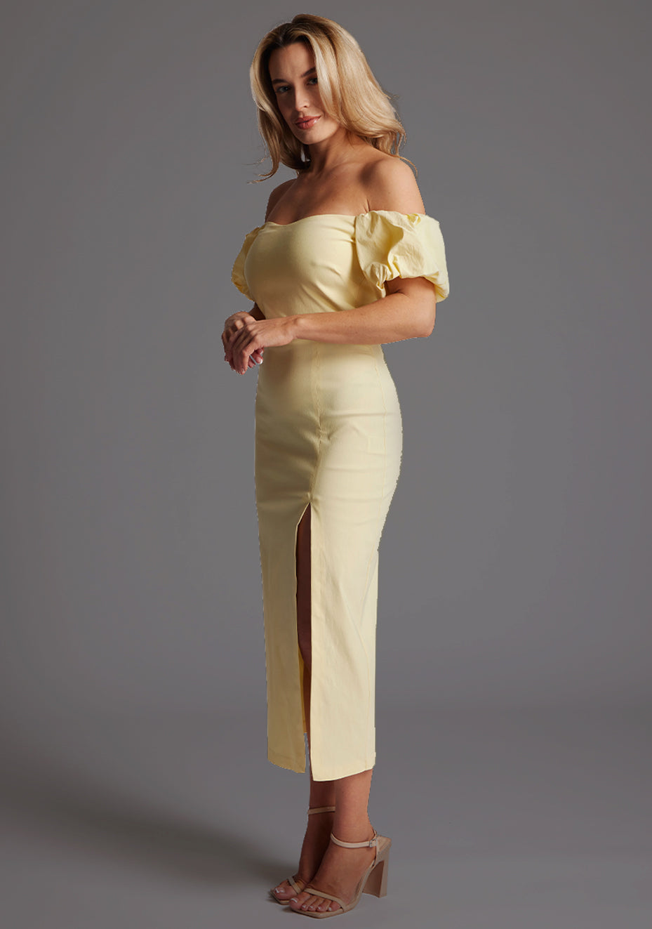 Front quarter image of a blonde model wearing a yellow bardot midaxi dress with a subtle sweetheart neckline, statement puff sleeves, a front skirt split and lace-up detail at the back. The design featured is the Vesper River Yellow Bardot Midaxi Dress