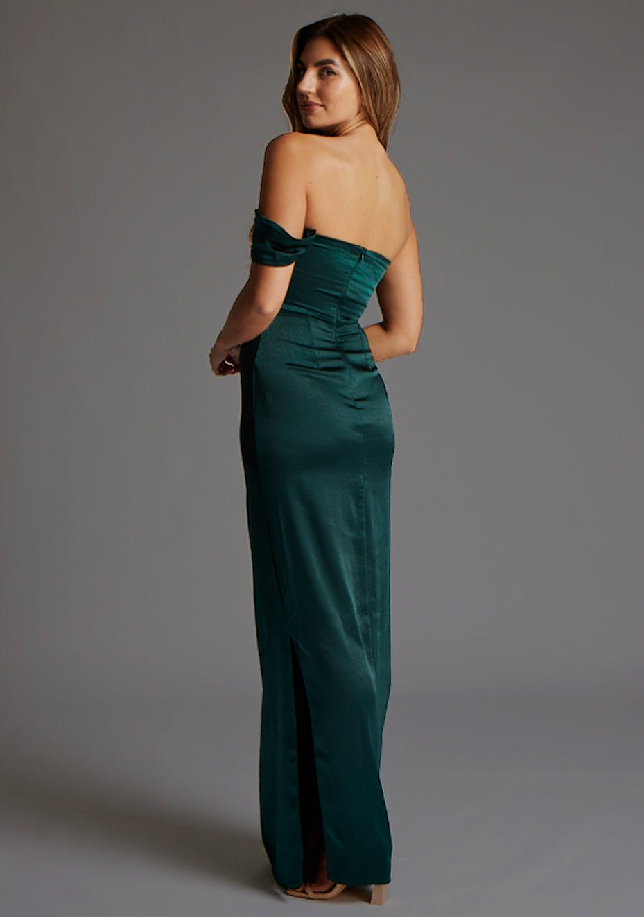 Front quarter image of blonde model wearing a pine green maxi dress with a one-sleeve off-shoulder design, with a satin fabric. The design featured is the Vesper Roselie Pine Maxi Dress