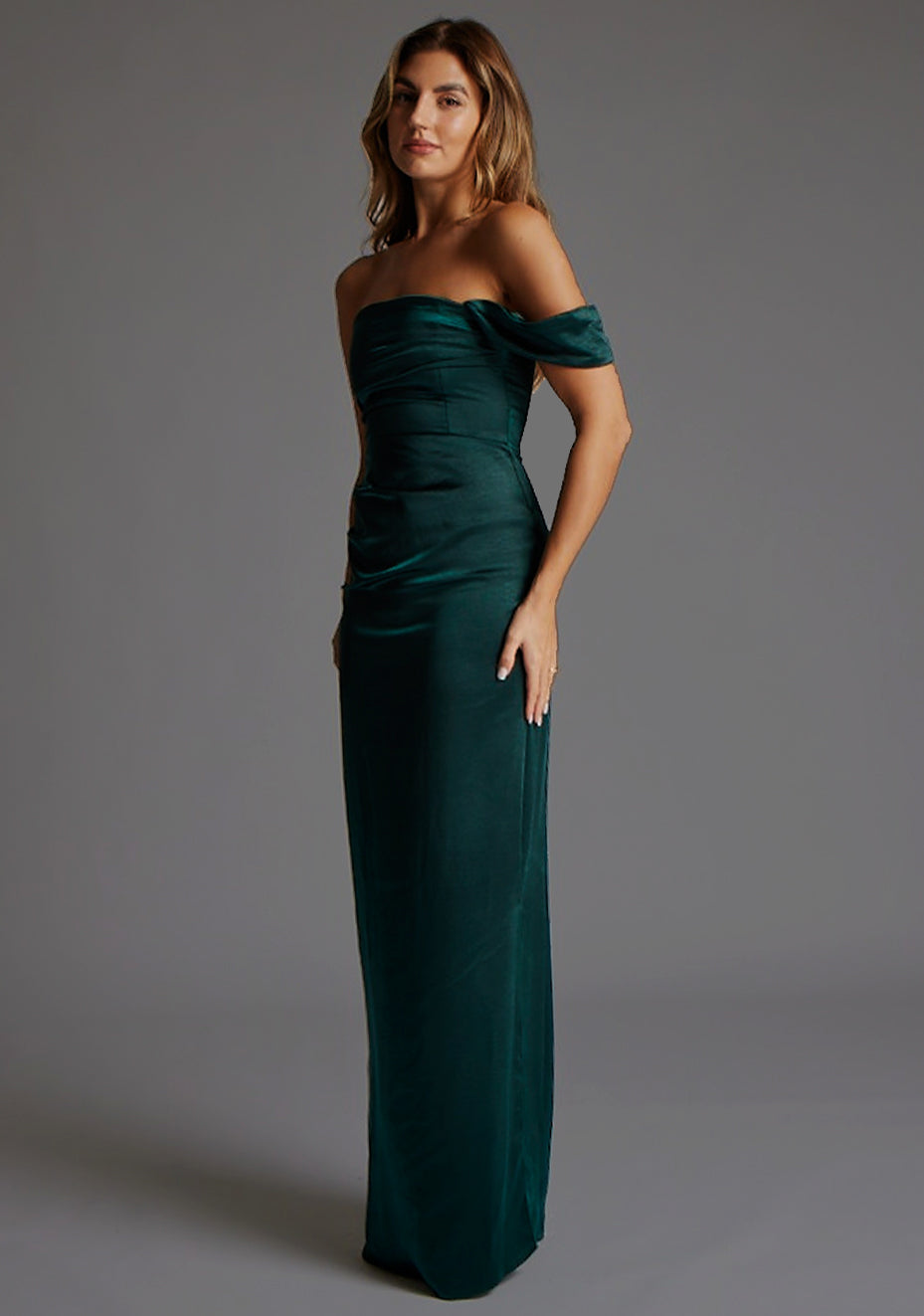 Front quarter image of blonde model wearing a pine green maxi dress with a one-sleeve off-shoulder design, with a satin fabric. The design featured is the Vesper Roselie Pine Maxi Dress 