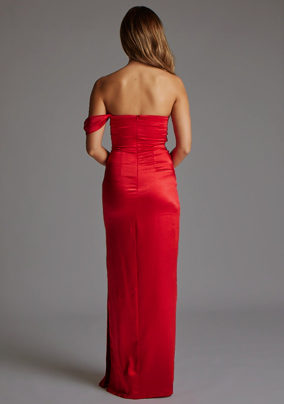 Back image of a blonde model wearing a Red Maxi Dress with a  one-sleeve off-shoulder design, crafted with luxurious satin fabric, a design features Vesper Roselie Red Maxi Dress