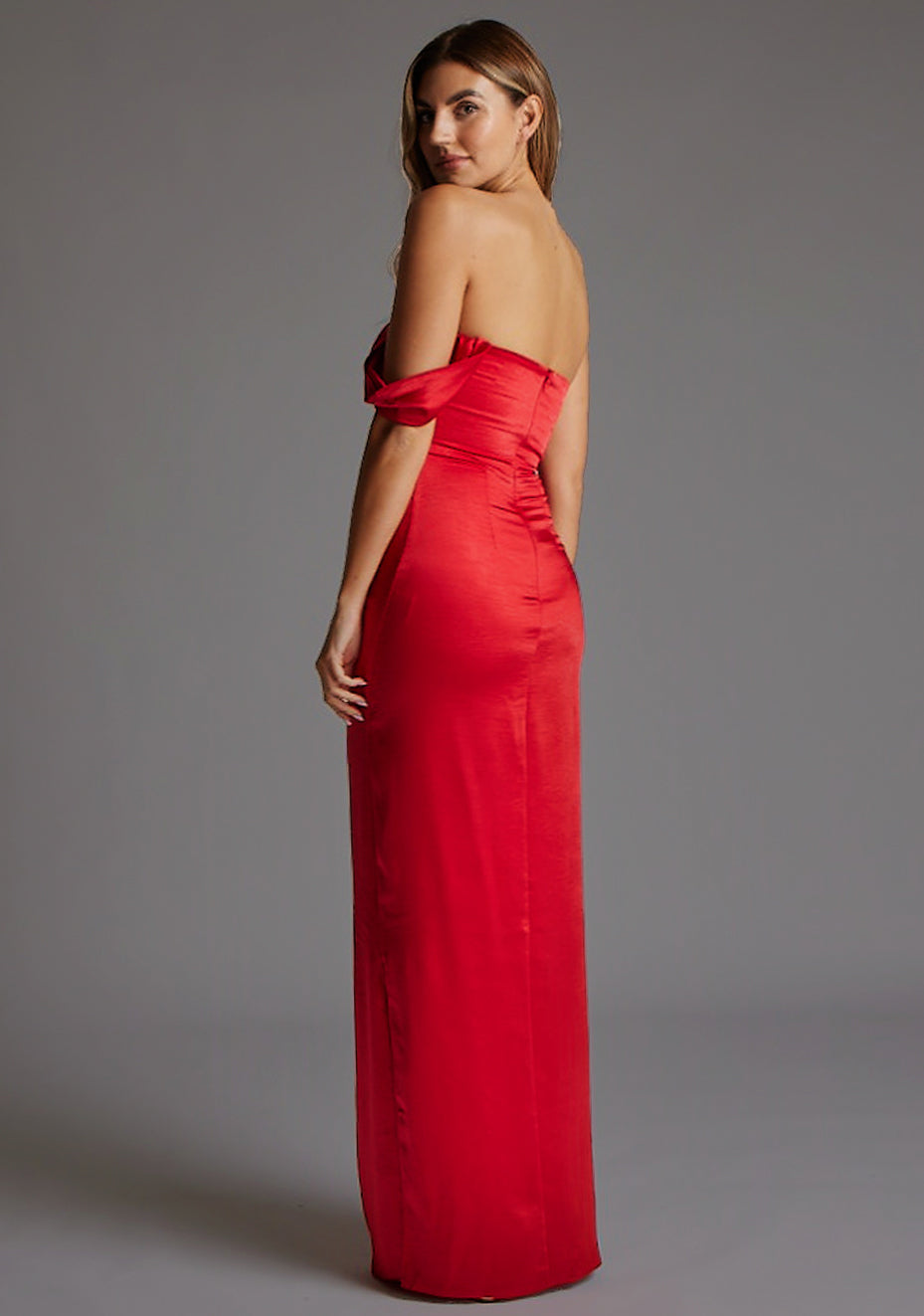 Back quarter image of a blonde model wearing a Red Maxi Dress with a  one-sleeve off-shoulder design, crafted with luxurious satin fabric, a design features Vesper Roselie Red Maxi Dress
