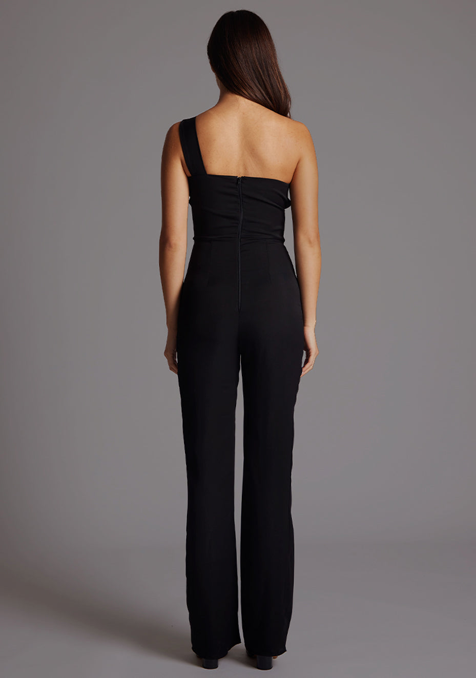 Back image of a  model wearing a black Jumpsuit with a stylish one-shoulder design, accentuated by a sophisticated front cut-out, slim fit and convenient back zip. The design featured is the Vesper Rosemary Black Jumpsuit.