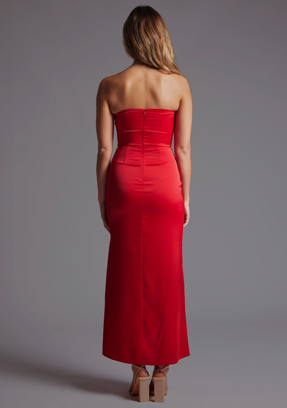 Back image of a blonde model wearing a red midaxi dress with a strapless design, front cutout with a sleek and slim fit silhouette. The design featured is the Vesper Sabine Red Midaxi Dress.