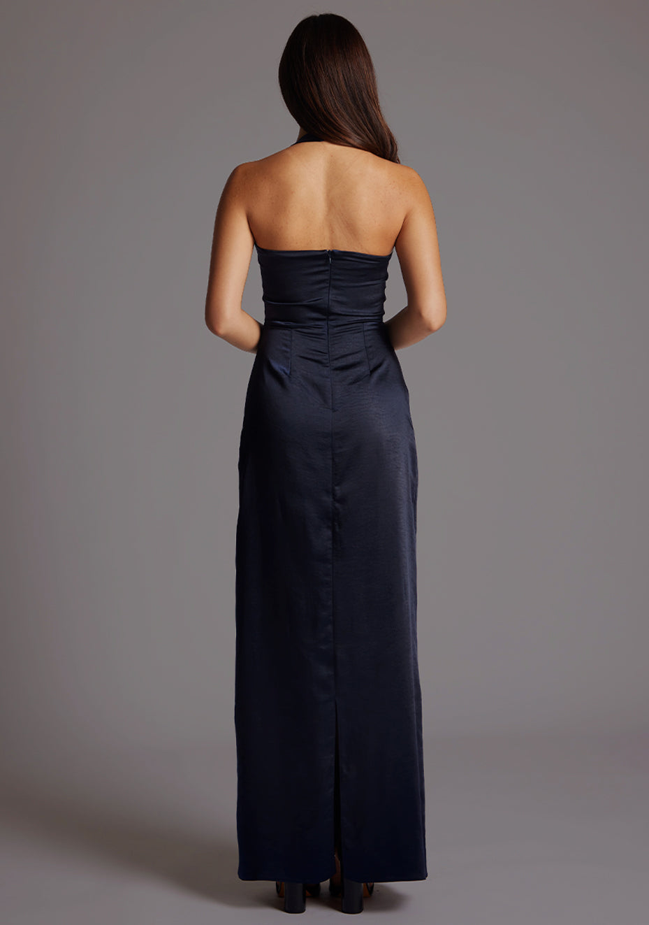 Back image of a model wearing a navy satin maxi dress with a halter cowl neck and low backless, the luxurious satin fabric slim-fit dress effortlessly flatters your figure.  The design featured is the Vesper Saige Navy Satin Maxi Dress.
