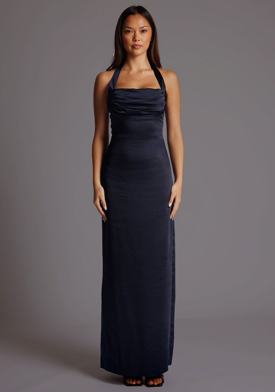 Front image of a model wearing a navy satin maxi dress with a halter cowl neck and low backless, the luxurious satin fabric slim-fit dress effortlessly flatters your figure.  The design featured is the Vesper Saige Navy Satin Maxi Dress.
