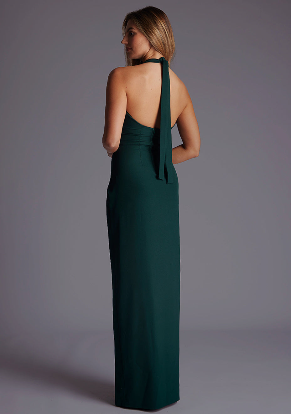 Back quarter image of a blonde model wearing a forest green maxi dress with a v-plunging neckline, halter design and a front skirt split. The design featured is the Vesper Sedona Forest Green Maxi Dress.