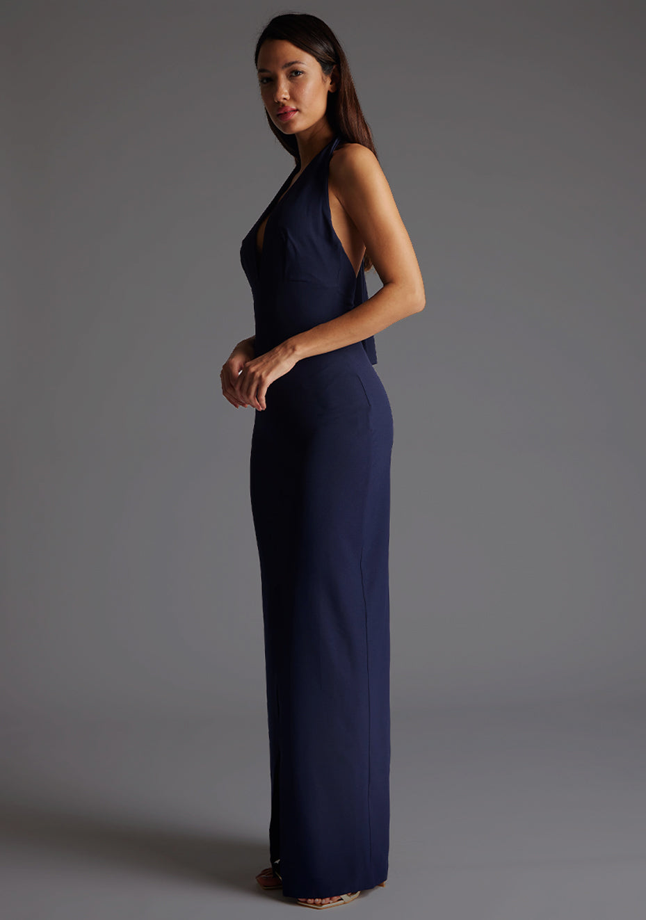 Front qaurter image of a blonde model wearing a navy maxidress with a v-plunging neckline, halter design and a front skirt split. The design featured is the Vesper Sedona Navy Maxi Dress