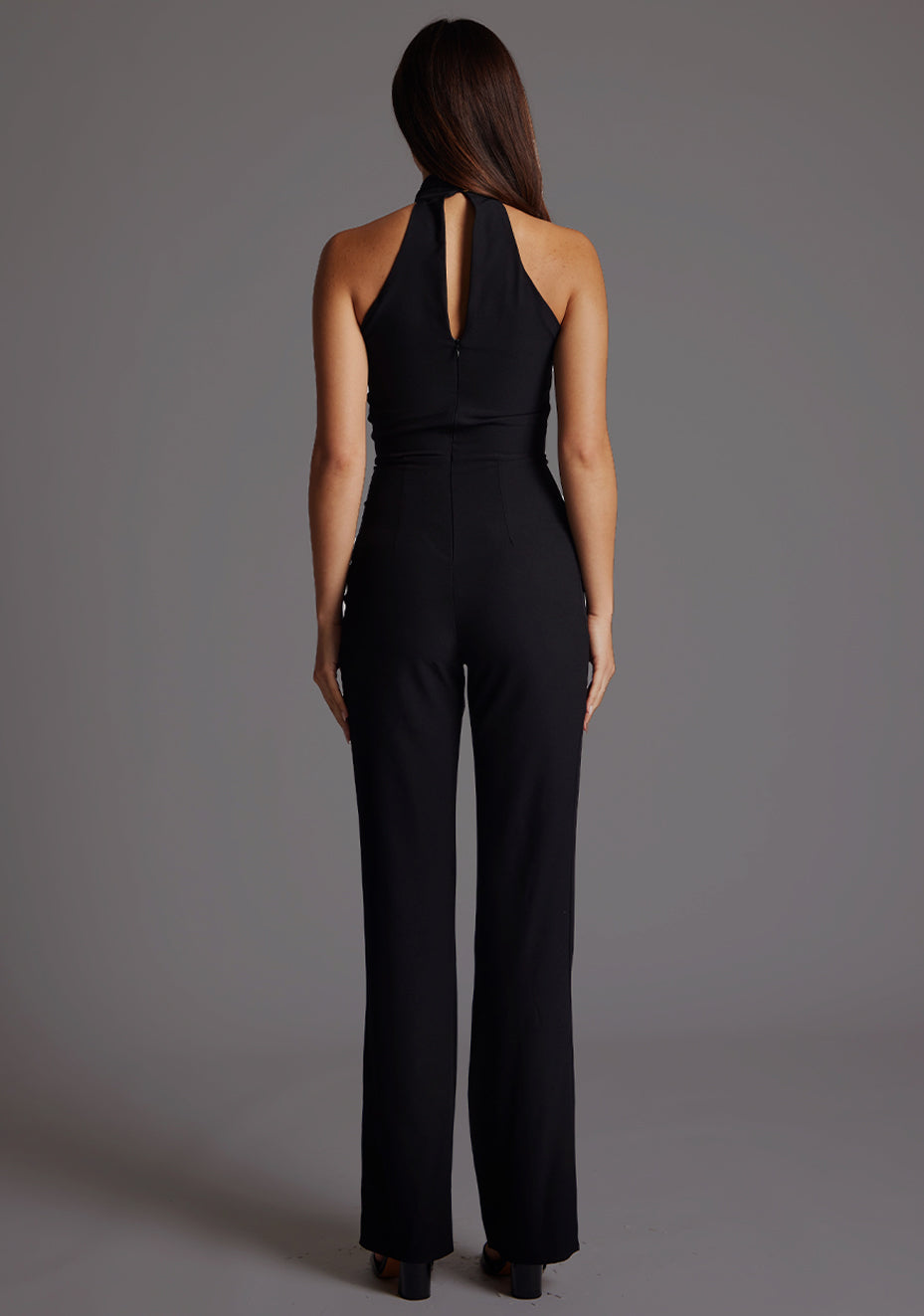 Back image of a model wearing a black jumpsuit with a high neckline, with a v-shaped cut-out at front bust, wide leg and invisible centre back zip. The design featured is the Vesper Stephanie Black Wide Leg Jumpsuit.