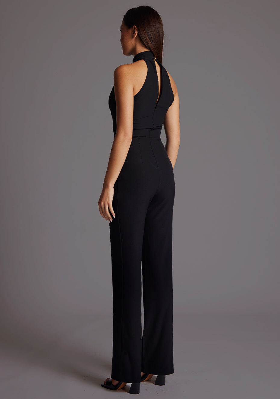 Back quarter  image of a model wearing a black jumpsuit with a high neckline, with a v-shaped cut-out at front bust, wide leg and invisible centre back zip. The design featured is the Vesper Stephanie Black Wide Leg Jumpsuit.