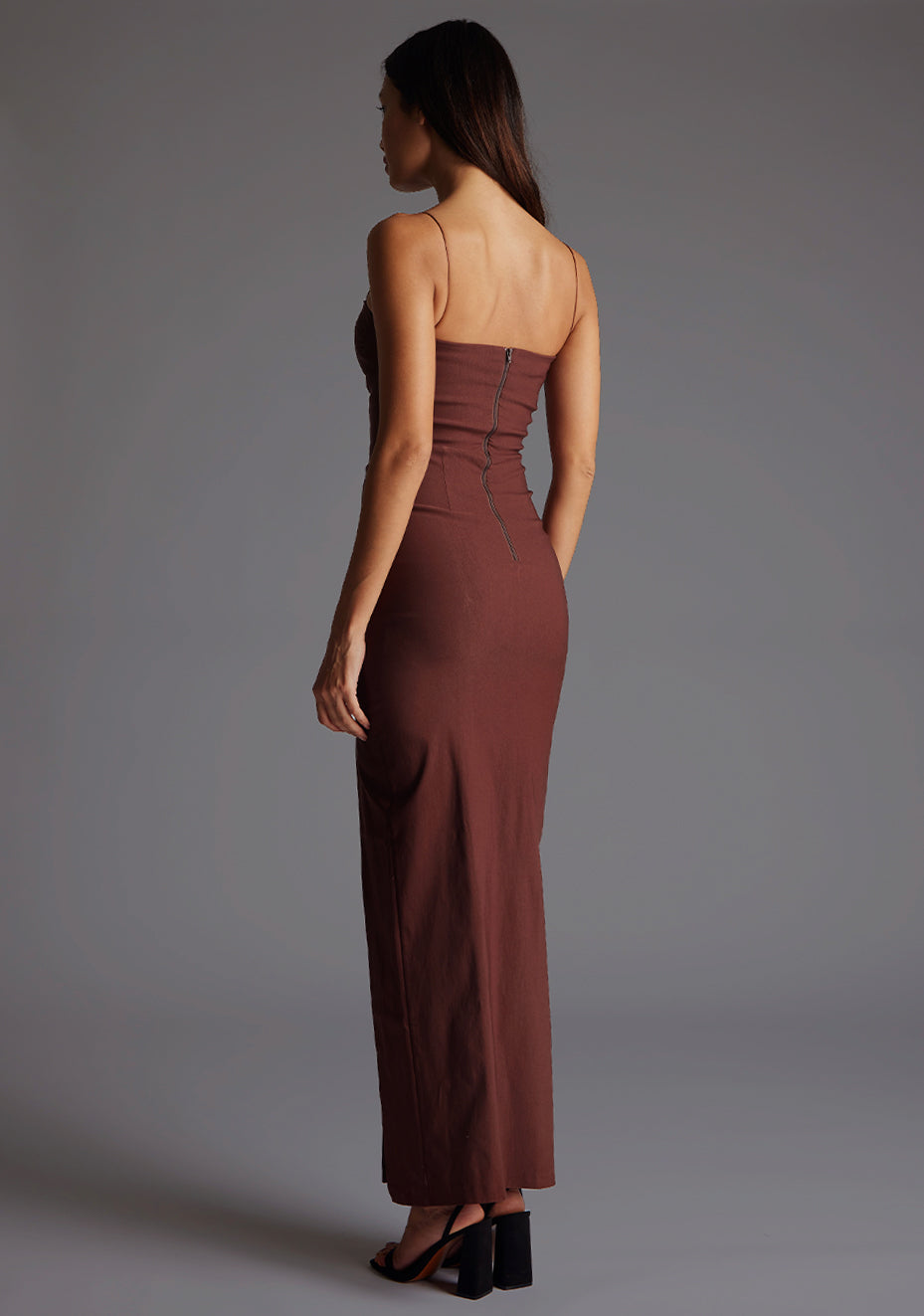 Back quarter image of a model wearing a chocolate maxi dress with a chic square neckline with an elegant cutout, complemented by delicate thin straps, back zip ensures a seamless fit and a front skirt split. The design featurd is the Vesper Suki Chocolate Maxi Dress.