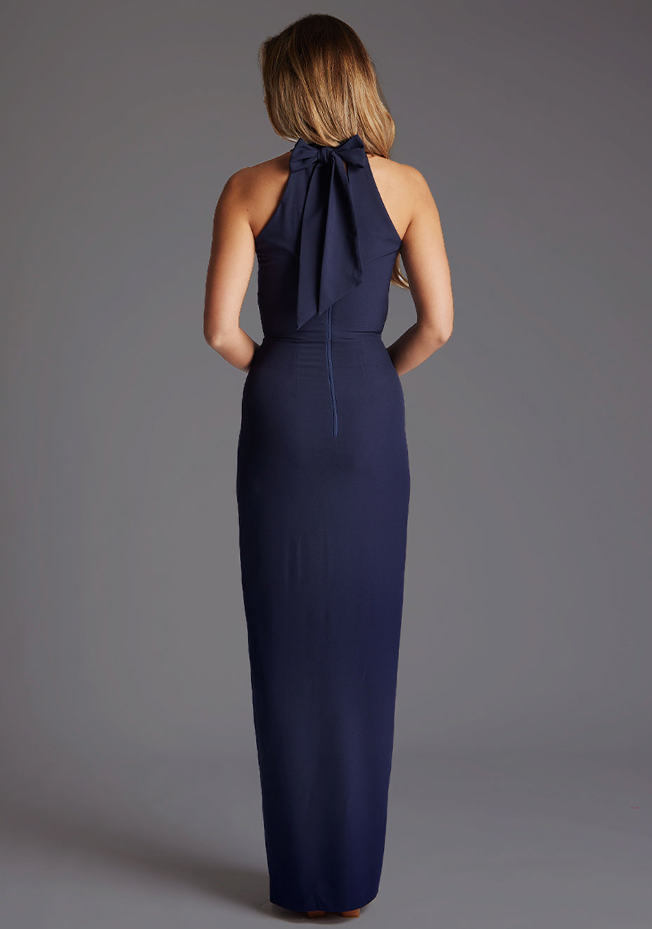 Back image of a model wearing a navy maxi dress with a halter neck design, front skirt split for added drama and  back zip for easy wear. The design featured is the Vesper Sydney Navy Maxi Dress.