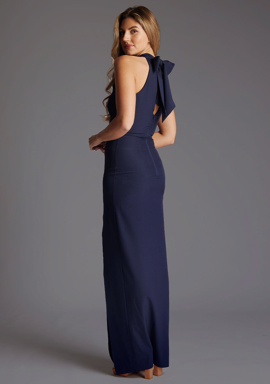 Back quarter image of a model wearing a navy maxi dress with a halter neck design, front skirt split for added drama and  back zip for easy wear. The design featured is the Vesper Sydney Navy Maxi Dress.