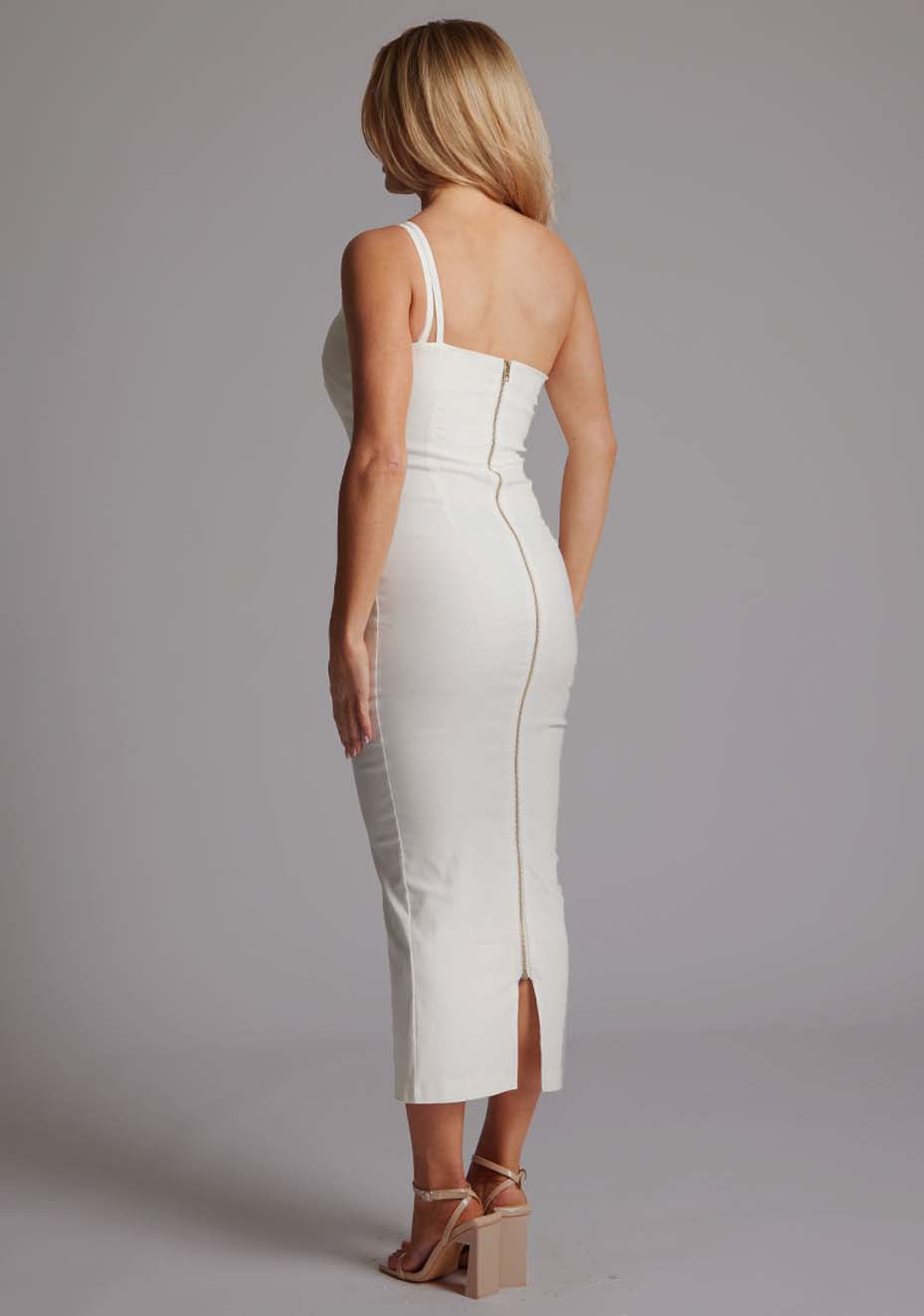 Back quarter image of blonde model wearing a ivory midaxi dress with an unique one-shoulder design, asymmetric neckline with a small cut-out and a bodycon fit, finished with a statement centre back gold zip. The design featured is the Vesper Tasha Ivory One Shoulder Midaxi Dress.