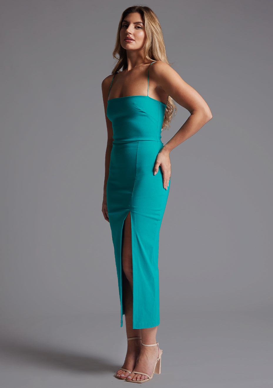 Front quarter image of a model wearing a peacock midaxi dress with a minimalistic strap details, front skirt split and centre back zip. The design featured is the Vesper Tate Peacock Midaxi Dress