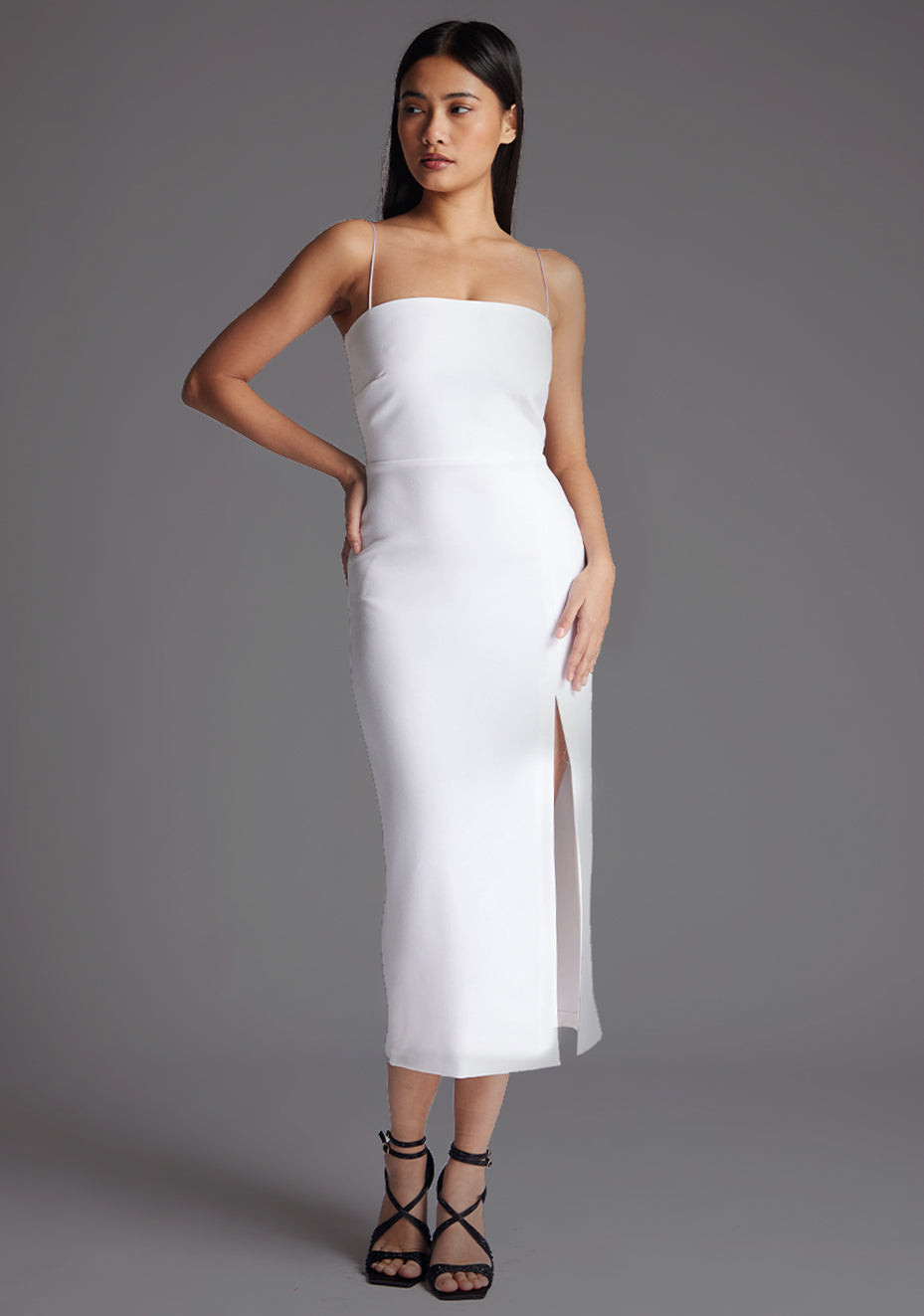 Front image of a model wearing a white midaxi dress with a minimalistic strap details, front skirt split and centre back zip. The design featured is the Vesper Tate White Midaxi Dress