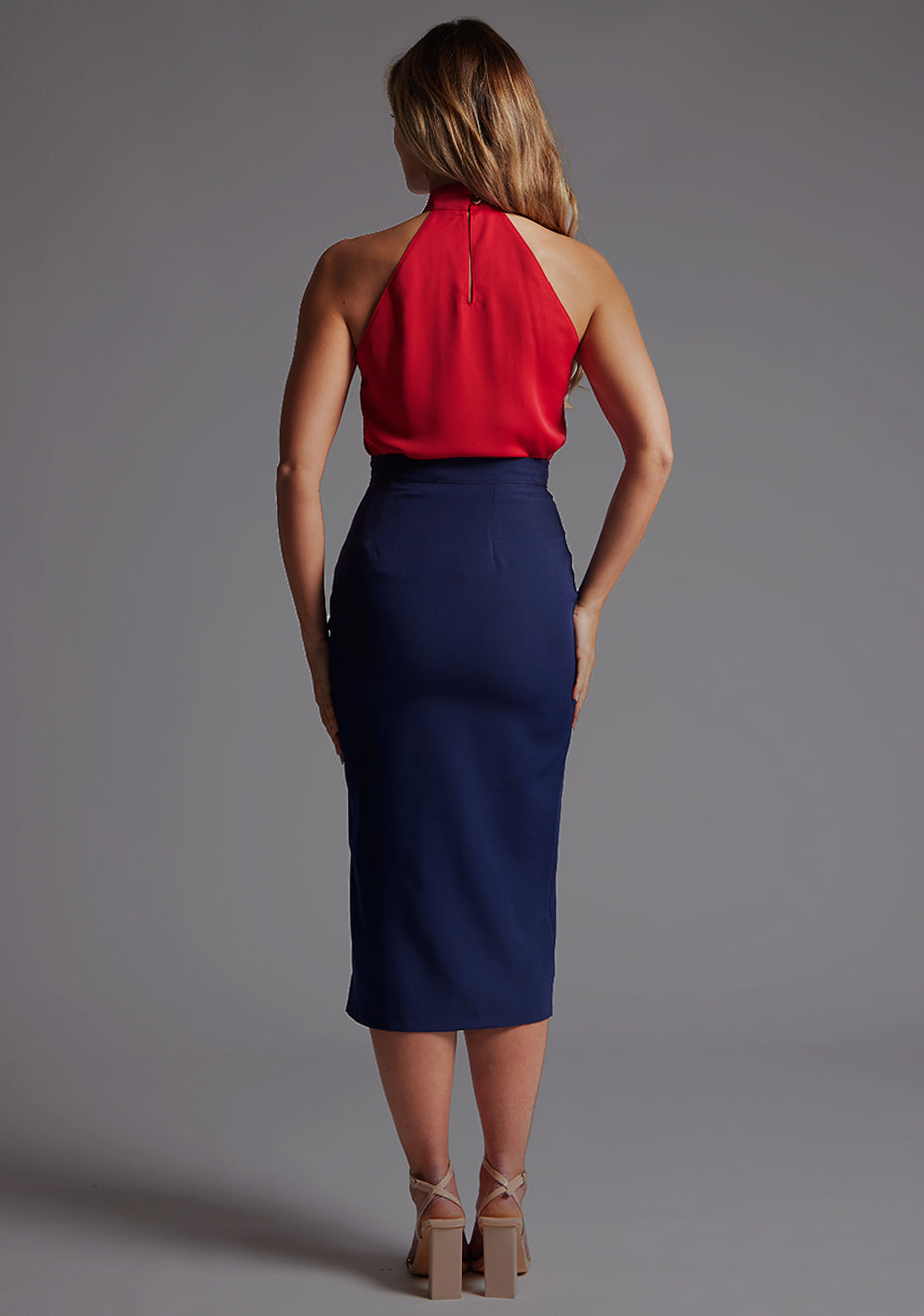 Back image of a  model wearing a red top with a high neck, sleeveless with wide pleat detail at front with a centre back cut-out with statement gold buttons fastening at the back of the neck. The design featured is the Vesper Toren Red Top pair with Vesper Tana Navy Fitted Skirt