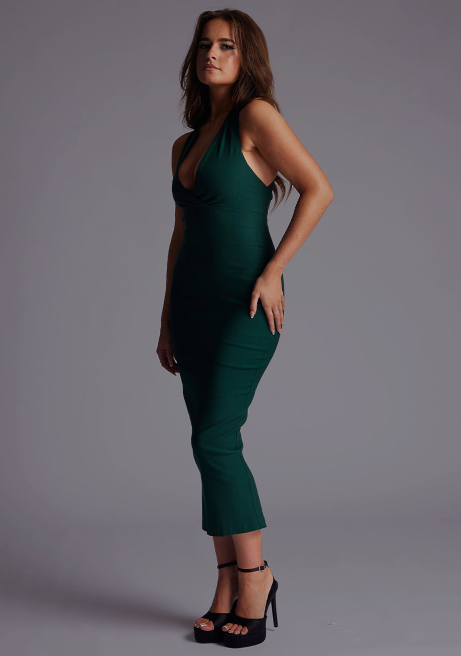 Front quarter image of a model wearing a forest green midaxi dress with a stunning sleeveless masterpiece, plunging v-neckline ,slim silhouette and cross back and gold zip at the back. The design featured is the Vesper Verona Forest Green Midaxi Dress