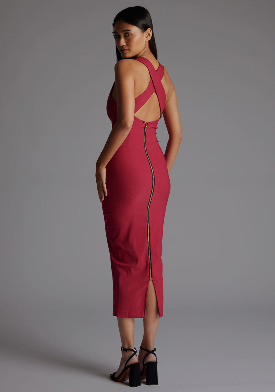 Back quarter image of a model wearing a raspberry midaxi dress with a stunning sleeveless masterpiece, plunging v-neckline ,slim silhouette and cross back and gold zip at the back. The design featured is the Vesper Verona Raspberry Midaxi Dress