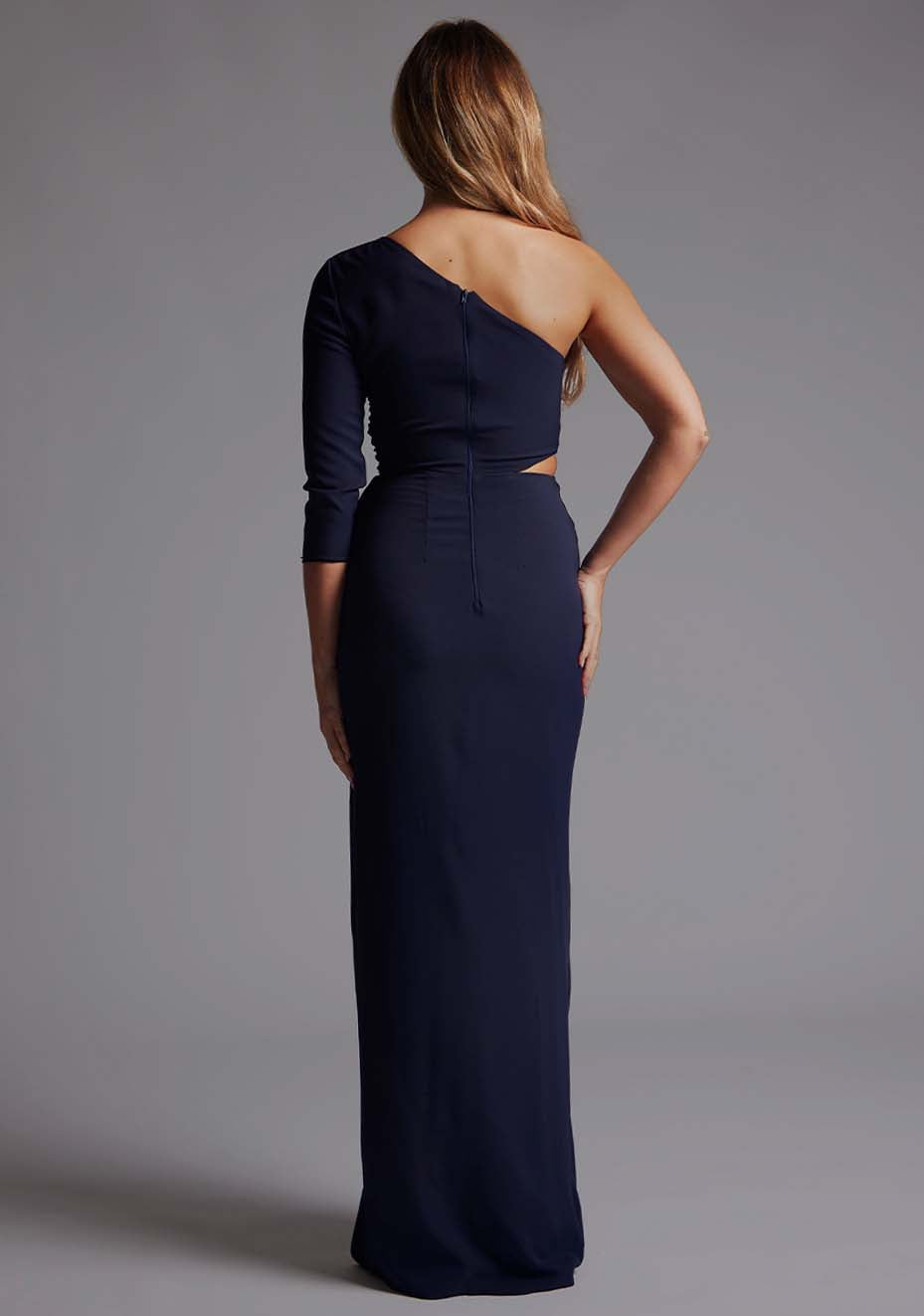 Back image of a blonde model wearing a  Navy Maxi Dress with an asymmetric long-sleeve design, coupled with a tasteful front cutout, creates a uniquely stylish statement. The design features is the Vesper Vienna Navy One Sleeve Maxi Dress
