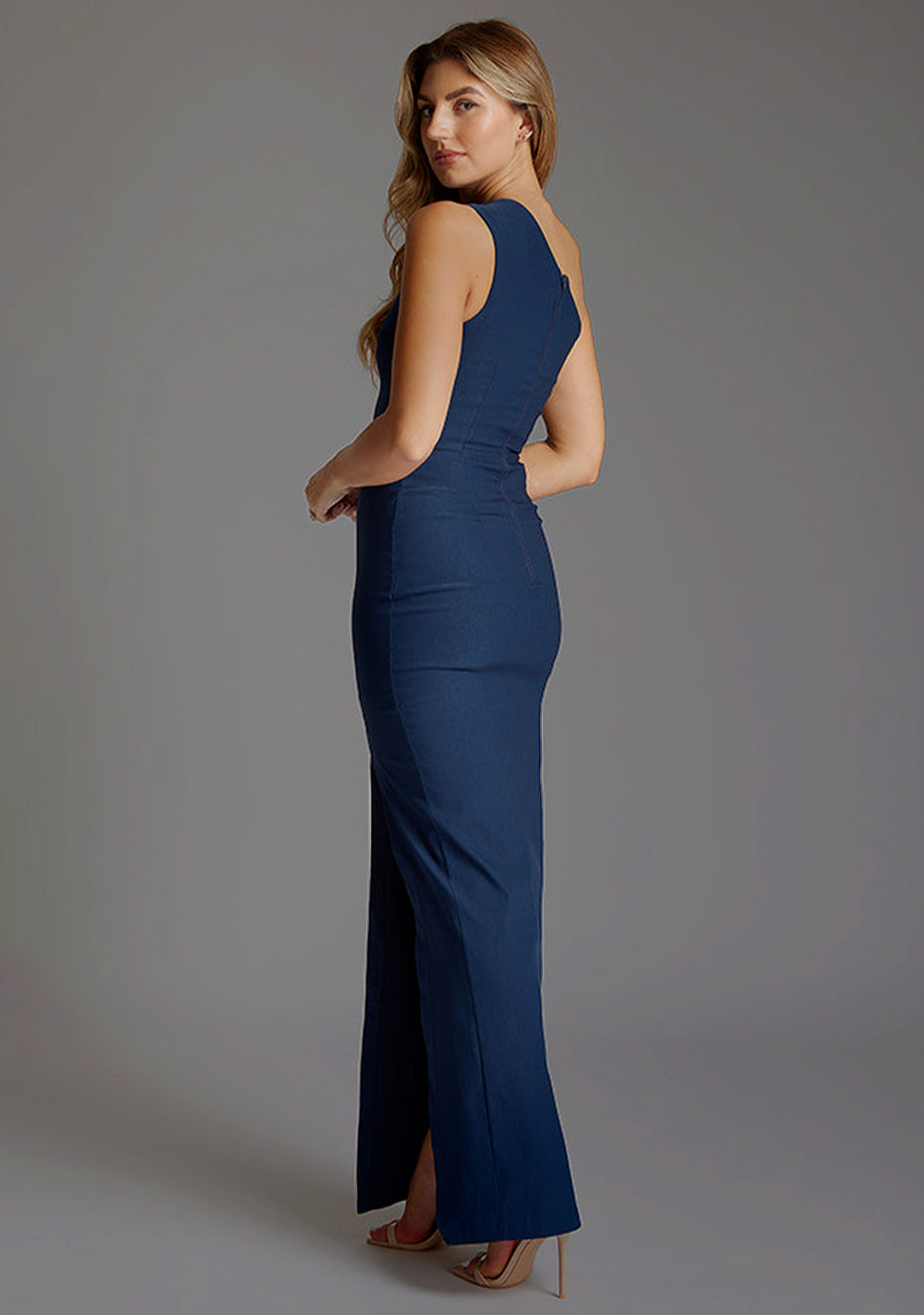 Back quarter image of blonde model wearing a Navy Maxi Dress with an asymmetric neckline with thigh-high leg split and cut out one shoulder design. The design featured is the Vesper Wren One Shoulder Navy Maxi Dress