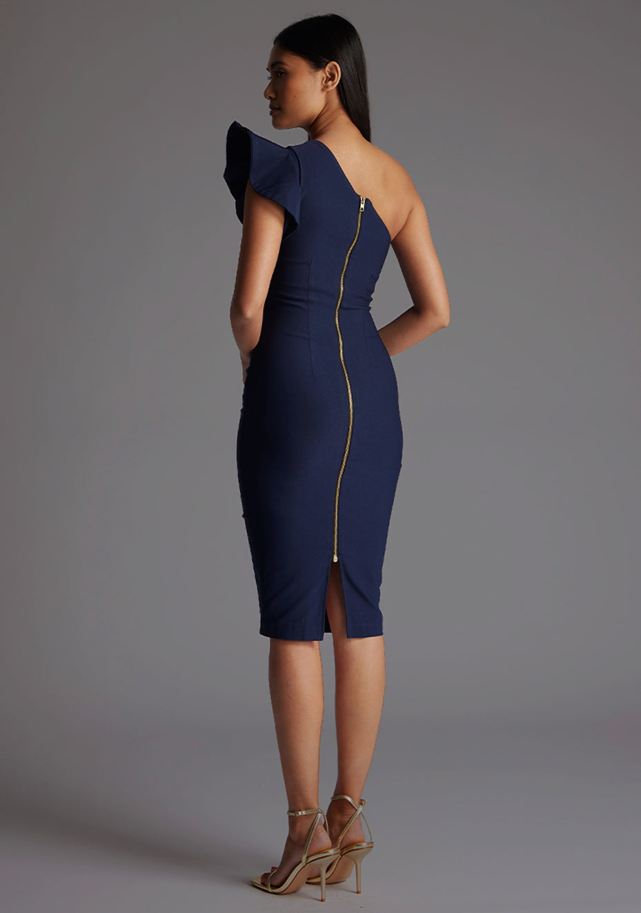 Back quarter image of a model wearing a  navy one shoulder midi dress with a stunning one-shoulder design with ruffle detail and an asymmetric neckline, all tied together with a gold centre back zip. The design featured is the Vesper Zanab Navy One Shoulder Midi Dress.
