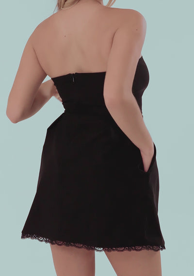 Model elegantly showcases the VSPR Emily Strapless Lace Trim Mini Dress in Black, twirling to highlight the flared skirt and delicate lace hem. The fitted strapless bodice enhances her silhouette as she walks confidently in silver crisscross heels. The minimal yet refined look is perfect for summer events, cocktail parties, or bridal occasions.