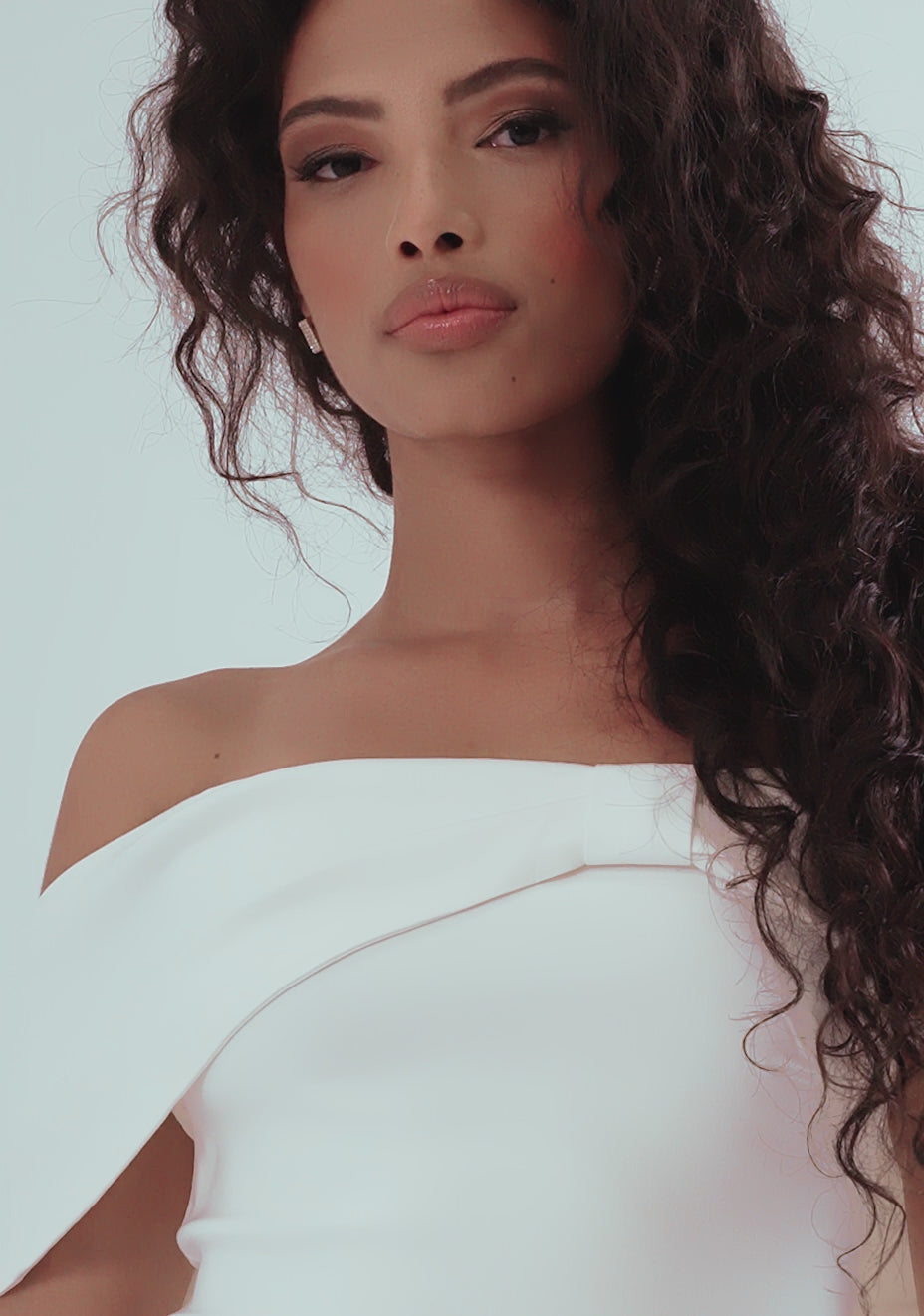 Model posing in the VSPR Sarah Bardot Bodycon Midaxi Dress in ivory, showcasing the flattering bardot neckline with ivory band across the arms, a striking bow detail, and a statement gold zip at the back; styled with a fascinator and heels for a polished, race-day-ready look.