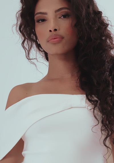 Model posing in the VSPR Sarah Bardot Bodycon Midaxi Dress in ivory, showcasing the flattering bardot neckline with ivory band across the arms, a striking bow detail, and a statement gold zip at the back; styled with a fascinator and heels for a polished, race-day-ready look.