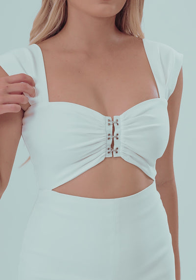 A model posing elegantly in the VSPR Morgan Ruched Bust Maxi Dress in Ivory. The dress features a sweetheart neckline, cap sleeves, and silver hook-and-eye closures on the bustier-style top. A cutout below the bust creates a chic two-piece illusion. The fitted high-waisted skirt falls to ankle length with a thigh-high slit. She pairs it with white strappy heels, creating a polished, modern look perfect for elegant evening events.