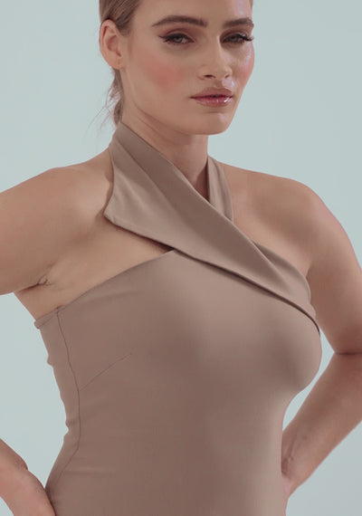 Model posing and turning in the VSPR Petra Taupe Midi Dress, showcasing the halterneck collar detail, button fastening at the back, and bodycon fit that hugs the figure. The soft, stretchy fabric moves gracefully as she poses, highlighting the elegant midi length and versatile taupe color in a city-inspired setting.