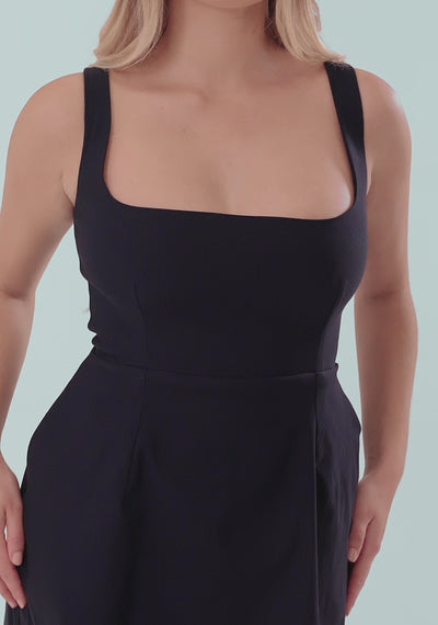 A model posing in the VSPR Camille Square Neck Mini Dress in Navy. The mini dress features thick shoulder straps and a square neckline, creating a sleek, classic silhouette. The skirt subtly flares for a playful, feminine touch. The model pairs the dress with silver high-heeled sandals with crisscross straps and ankle closures, completing an elegant look suitable for cocktail parties or evening events.